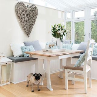 a dining table with wooden top and white legs in front of a built in bench seat and dining chair, in a light airy conservatory, with a large heart wooden twig decoration on the wall