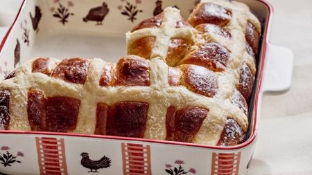 A square ceramic baking dish with hand painted red stripes and chicken motifs around it, filled with hot cross bins, on a white tablecloth. 
