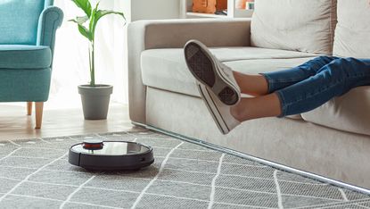 A woman rests on the sofa as a robot vacuum cleans the carpet