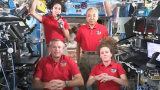 four astronauts in red shirts pose for a video message in a space station full of electronic equipment and computers