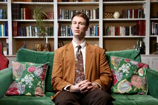 Hugo Bolton as Tony in AIDS: The Unheard Tapes. He is wearing a rust-coloured blazer over a white shirt with a paisley tie and black pinstripe trousers, sitting on a green sofa and looking into the camera. There are shelves full of books and plants on the wall behind him.
