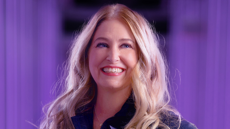 A white woman with blonde hair smiles and stares up a to the right.