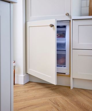 A kitchen with cream kitchen cabinets with one opening up to a freezer, two drawers, and a light brown wooden floor