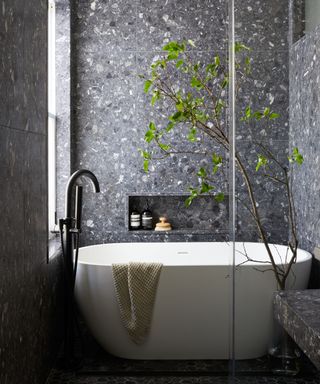 A small bathroom drenched in gray terrazzo tiles with a small white freestanding bathtub, niche wall storage, and a black faucet