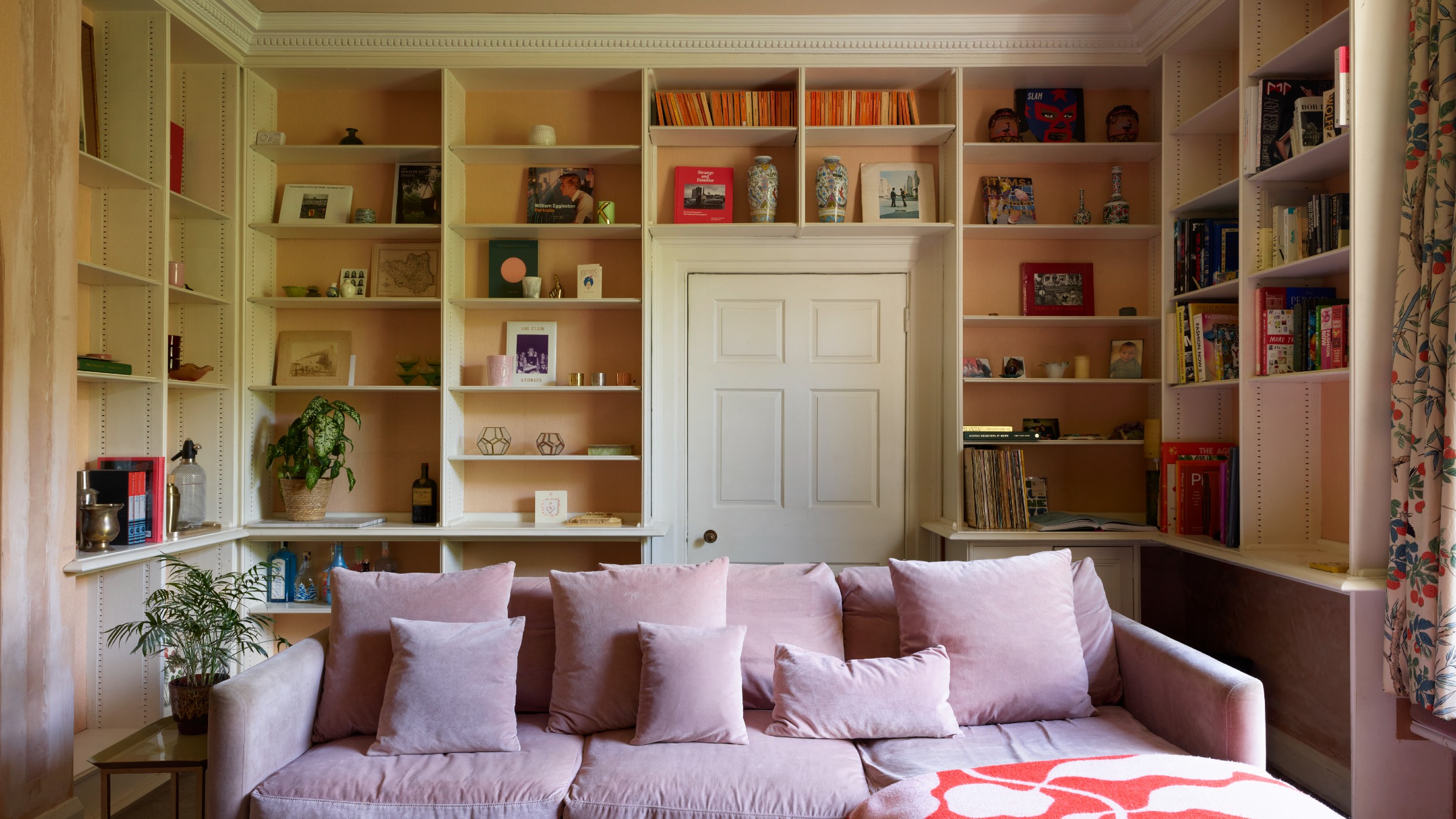 A living room with a pink velvet sofa and a built-in bookshelf covering three walls and surrounding a door 