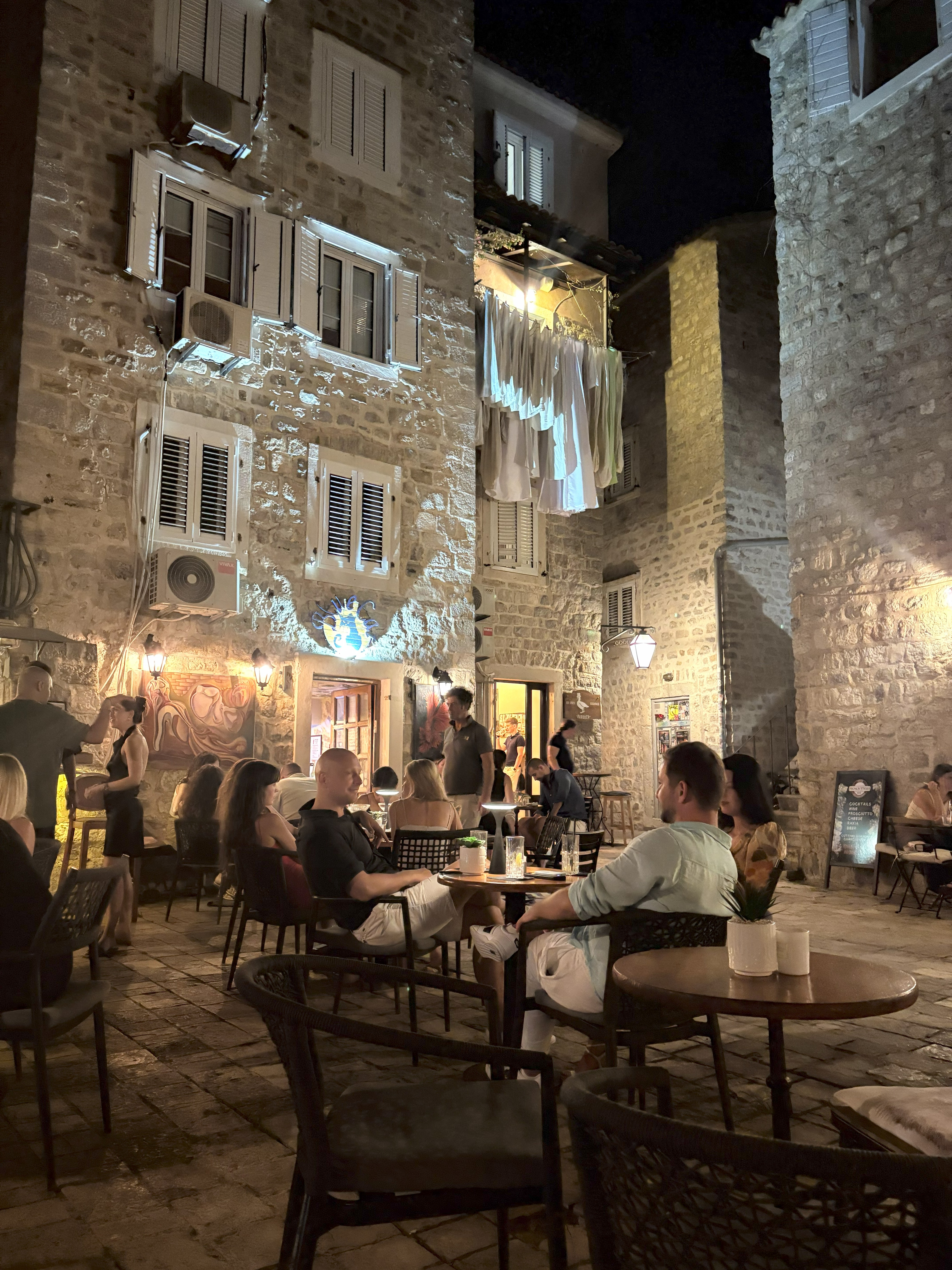 A rocky town's square features round wooden tables, laundry hanging from its balconies, and a warmly suffused atmosphere.