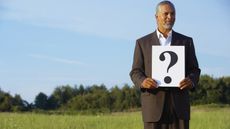 An older man stands in a field and holds a sign that has a question mark on it.