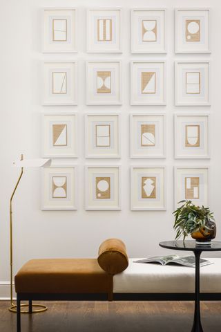 gallery display of framed artwork on white wall with bench chaise in front and reading light with dark metal wine table