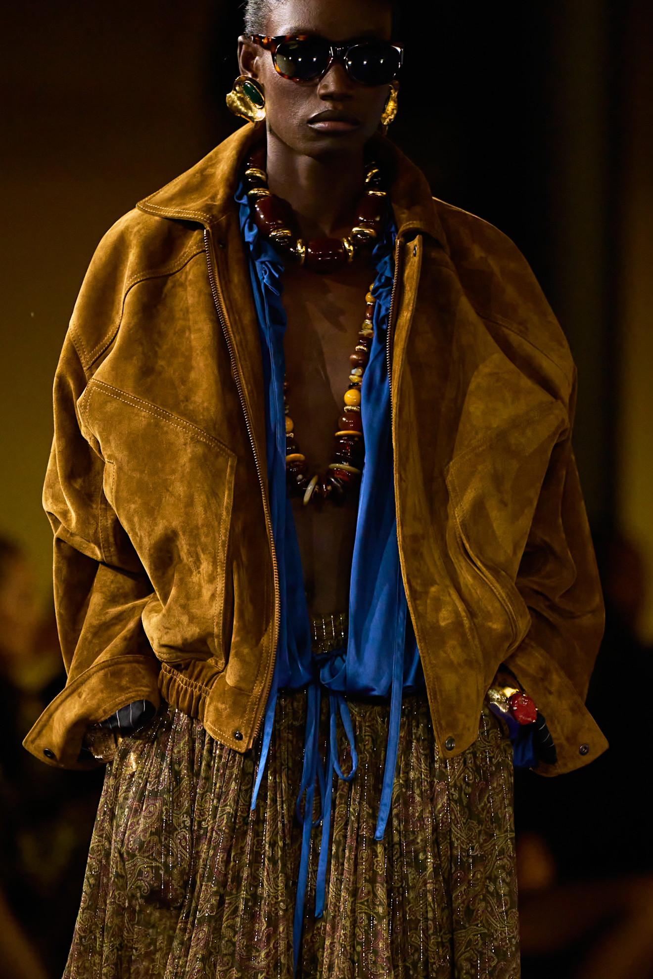 Saint Laurent runway model wearing a large beaded necklace, a tan jacket, and a loose brown skirt.