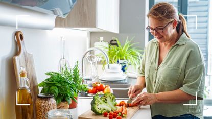 Woman standing over chopping board doing meal preparation of fruits and vegetables with sunlight coming through window in front of her, representing how much weight can I lose in a month in a healthy way