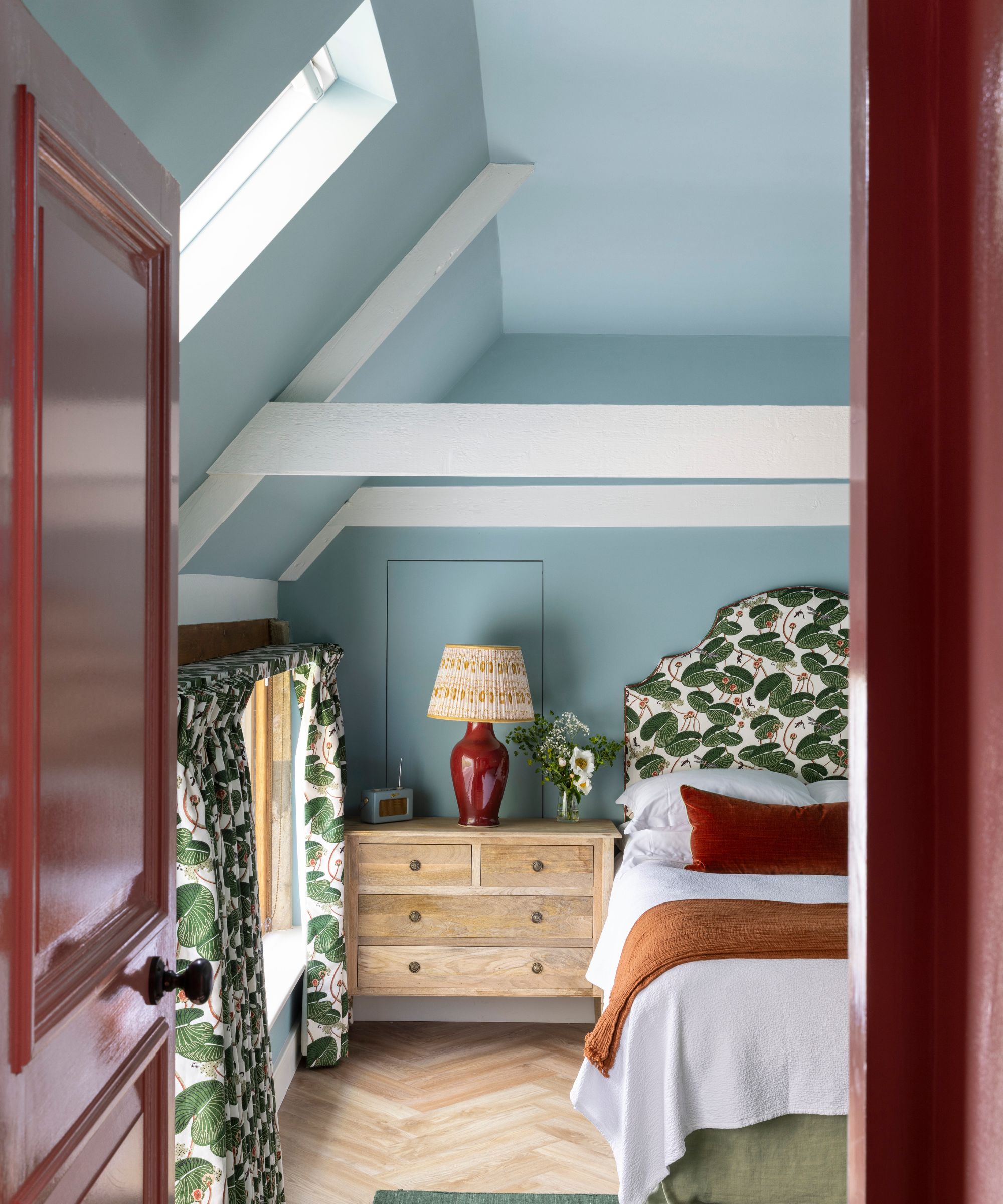 Bedroom with pale blue walls, red door, floral curtains and matching headboard, and wooden side table with lacquer red lamp