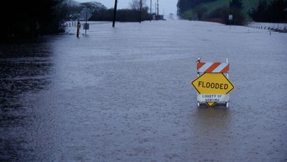 A warning sign saying "flooded" in the middle of a flooded street in the rain.