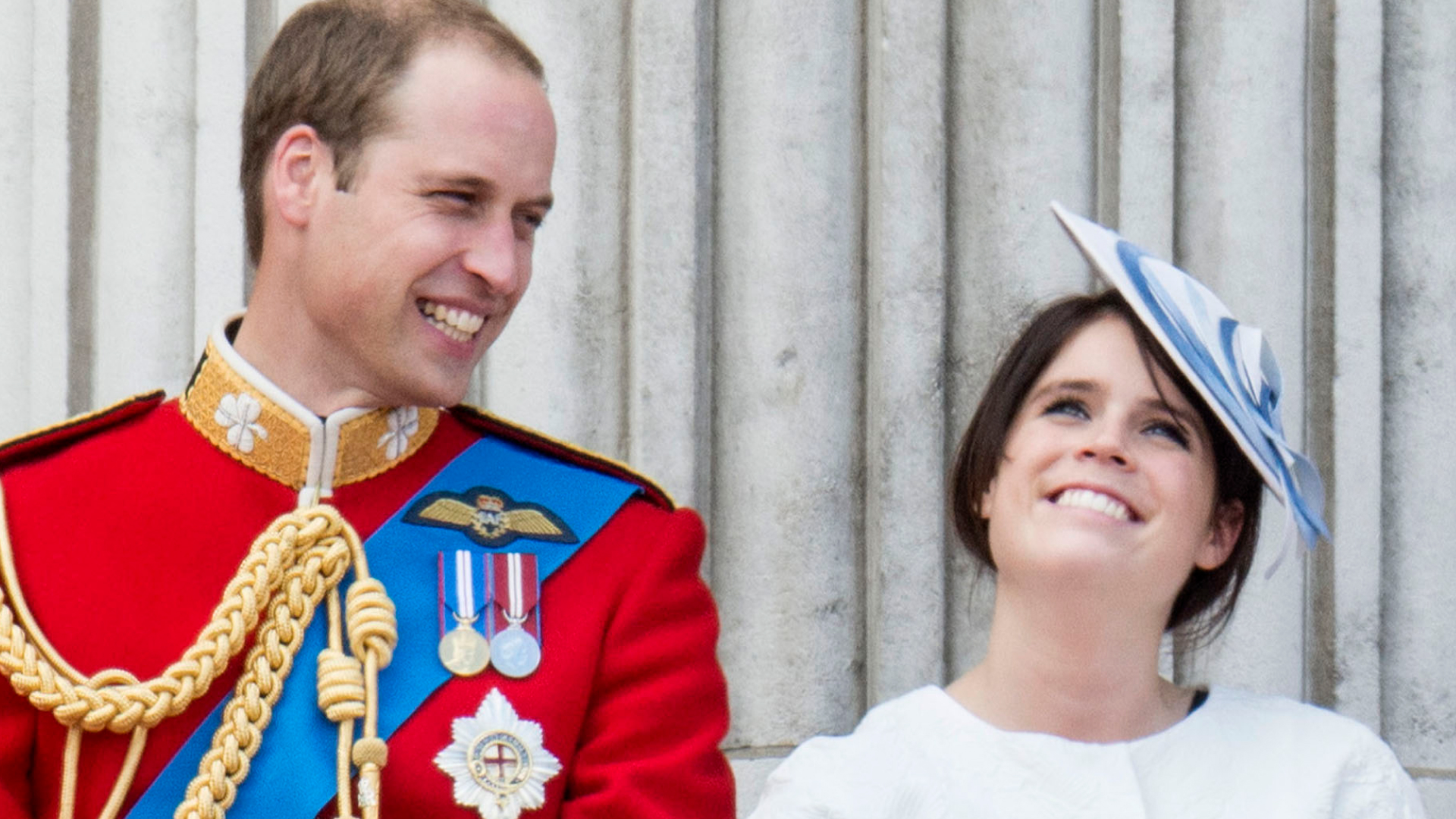 Prince William laughs while wearing red military uniform as he stands next to Princess Eugenie, who is wearing a white dress and a blue and white hat on the Buckingham Palace balcony