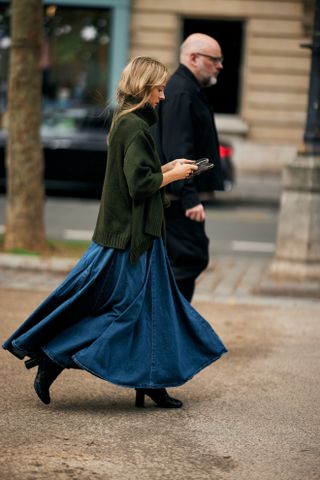 A woman at Paris Fashion Week Spring 2025 with her blond hair tucked into a green turtleneck and wearing a blue denim maxi skirt