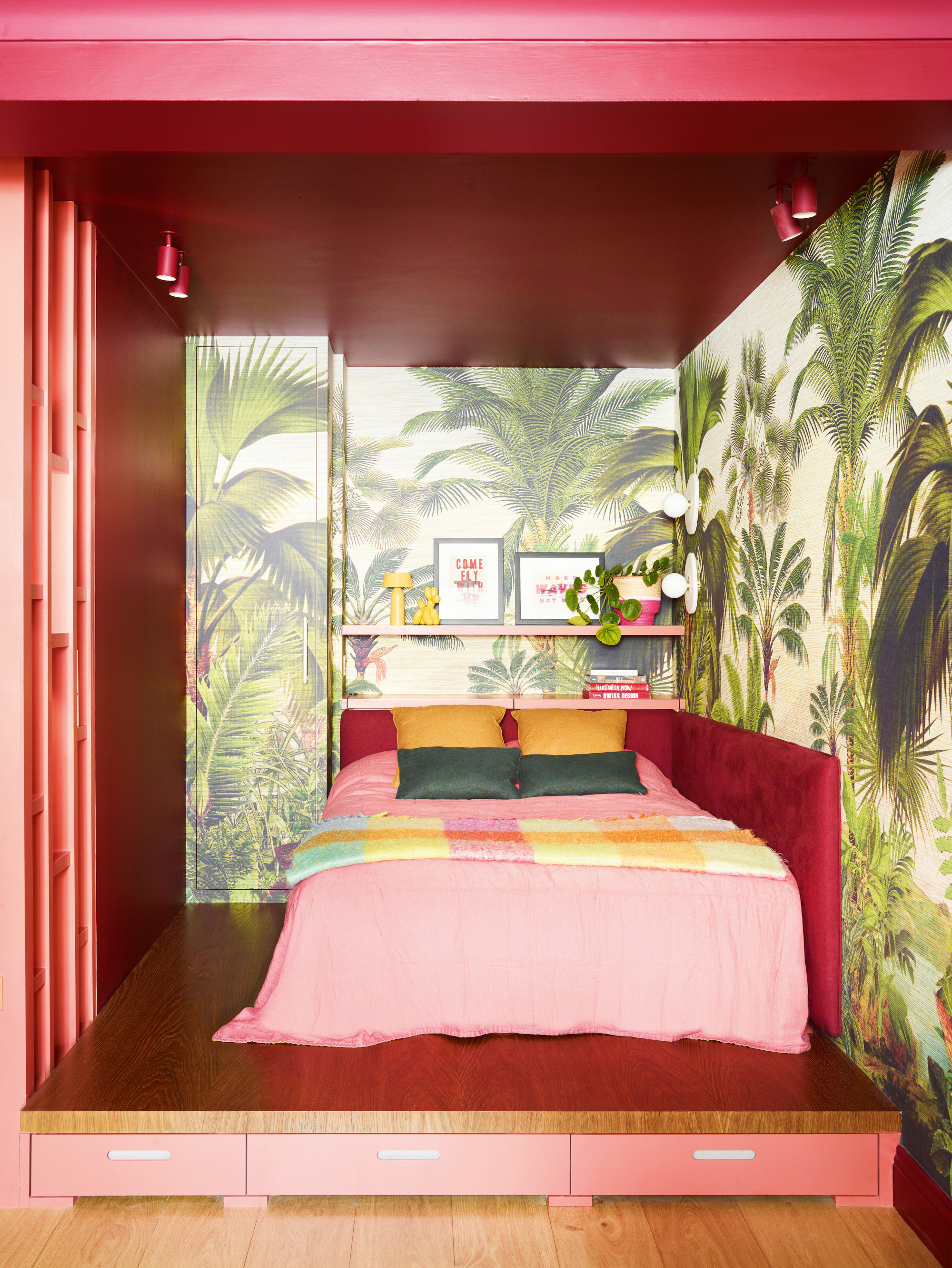 Bed on raised platform with velvet headboard, green palm tree wallpaper, dark pink walls and ceiling, pale pink bedding and green, yellow and pink throw