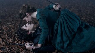 Emily Bader as Lady Jane Grey laying on top of Edward Bluemel as Guildford Dudley, they're both holding daggers.