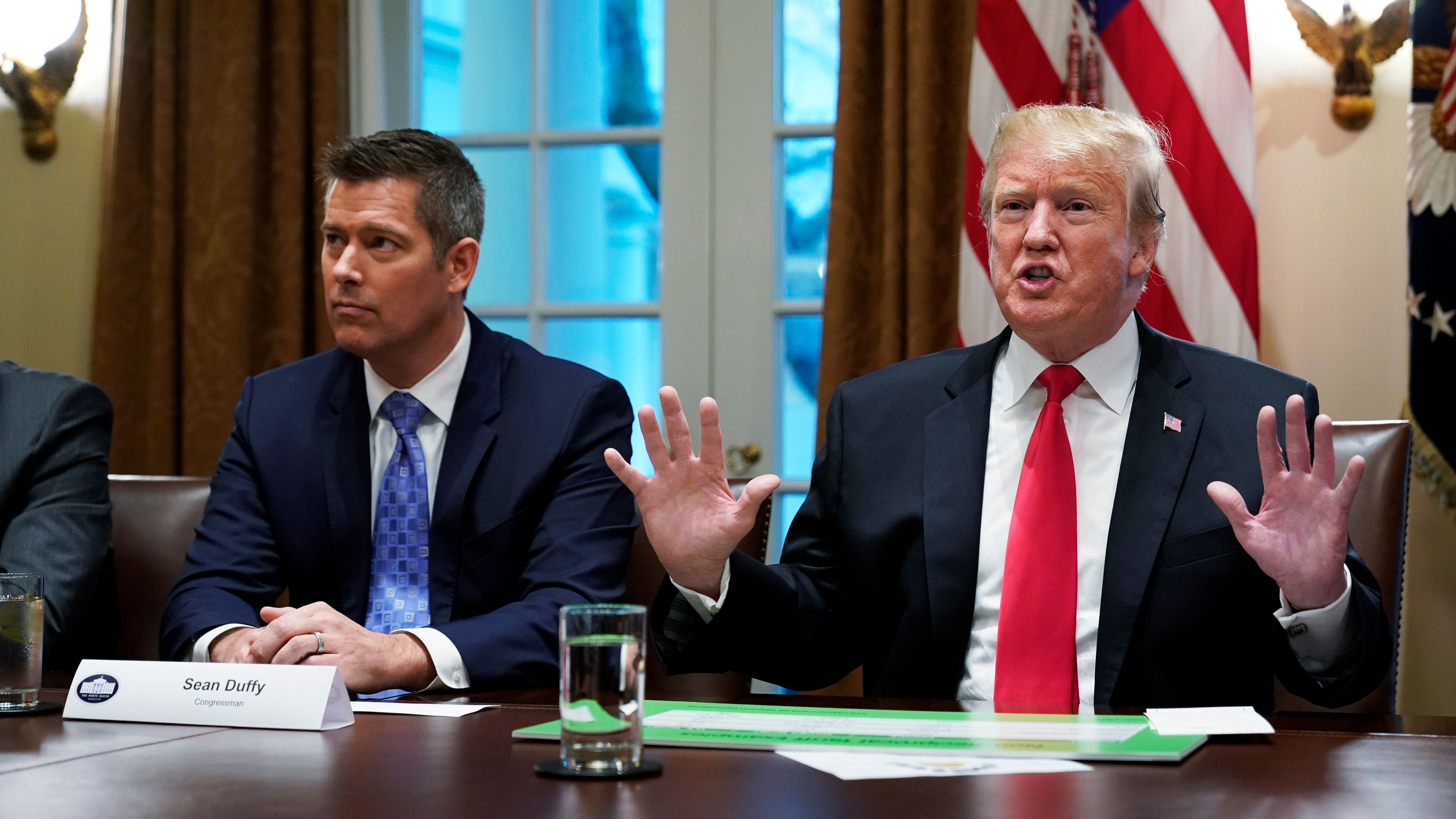 Two men in dark suits sit at a brown wooden table with a single glass of water. The man on the right has his hands raised and his lips pursed.