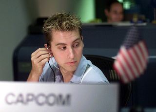 lance bass wears a headset while sitting down in an office chair. in front of him, blurry, is a sign with the word "capcom" and a small american flag