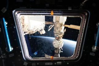 a window view of the space station. the curve of earth is visible in the background. a line of modules is attached to one another on the right-hand side of the picture, with two solar panels visible on either side. on the left side of the picture is a block-shaped module partially obscured by the window frame