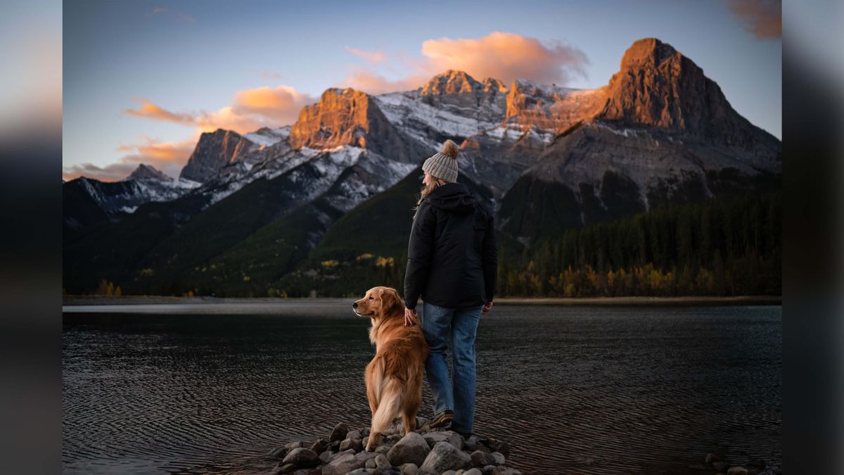 Dog and owner standing on rock pile at lake shore against backdrop of rocky mountains.