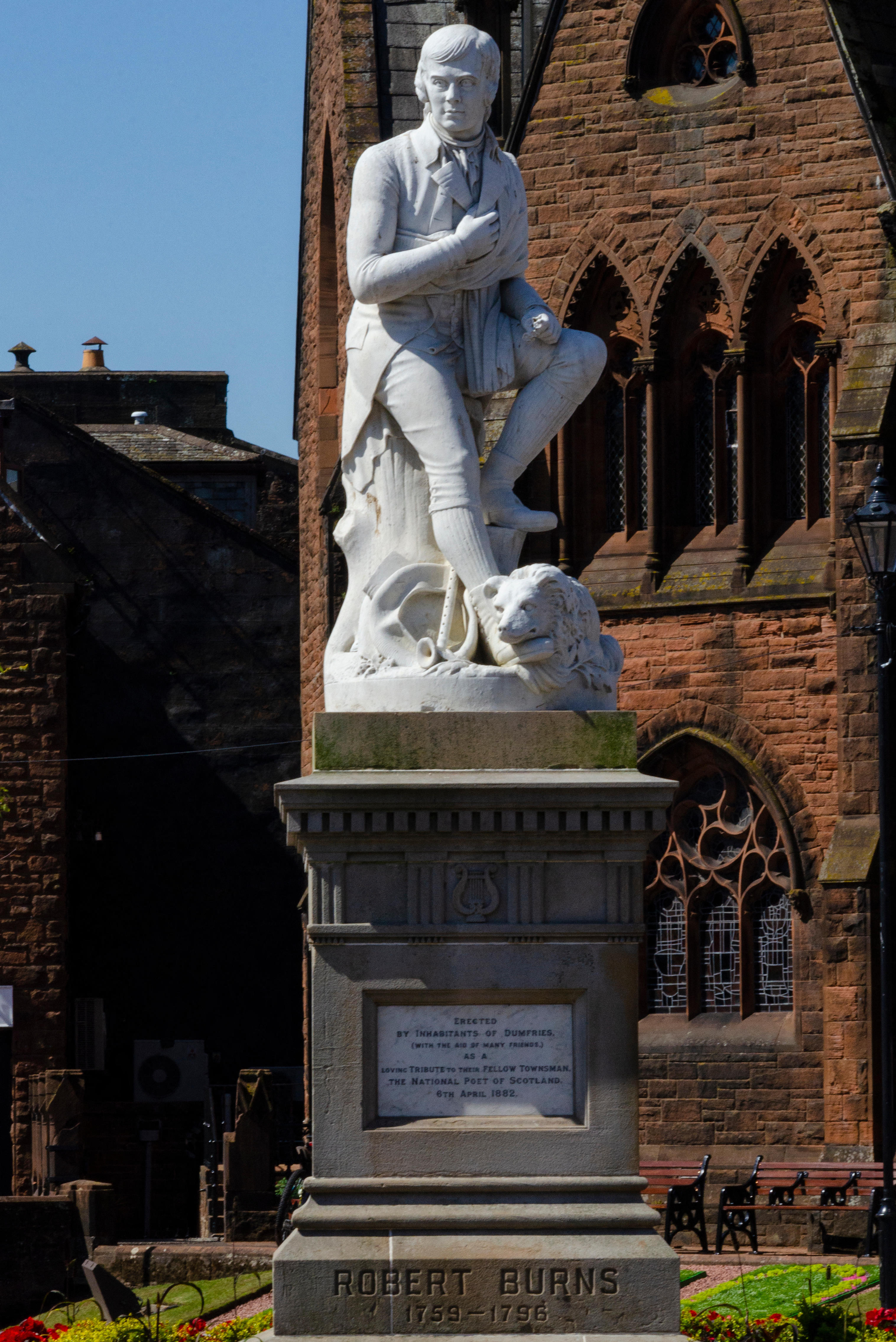 Statue of Robert Burns in Dumfries, standing with a dog at his side on a stone pedestal, with a red sandstone church in the background.