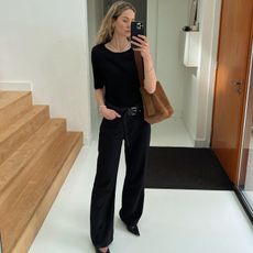 Fashion influencer Anouk Yve taking a mirror selfie wearing a chic fall outfit with belted jeans, black pointed heels, a short-sleeve black knit, and a suede bag.