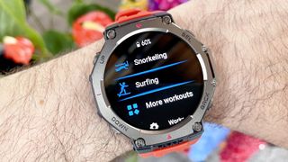 The Amazfit T-Rex 3 shown close-up on a user's wrist with the snorkeling and surfing workout tracking modes displayed; colorful flowers are out of focus in the background