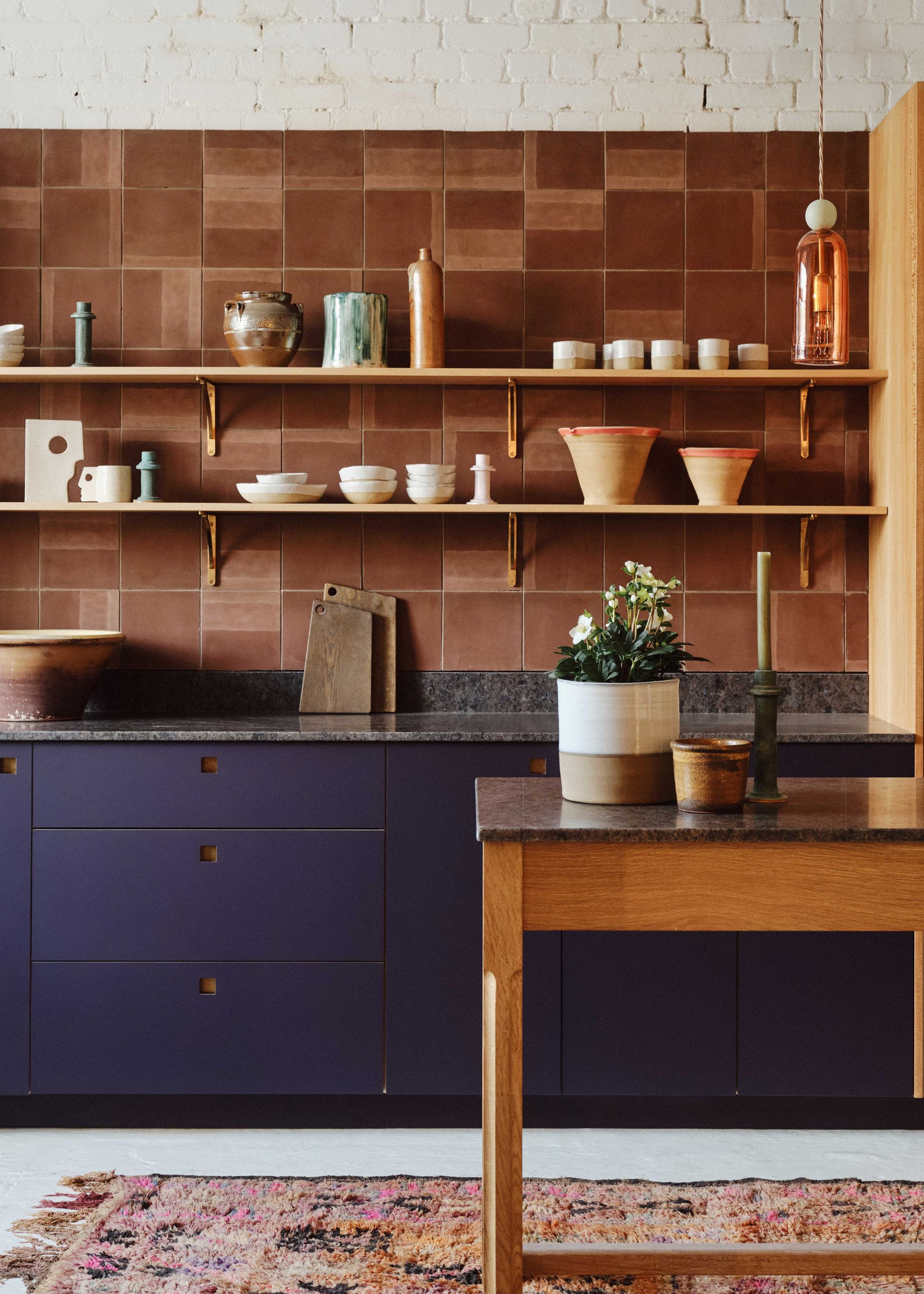 A kitchen with a table in the middle. On the table is a potted plant as well as a candle and candle holder. There is also space for open shelving in the kitchen with lots of bowls, pots and other kitchen essentials.