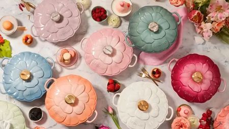 A set of Le Creuset brasiers with petal shaped lids on a marble counter surrounded by flowers, macarons, and strawberries