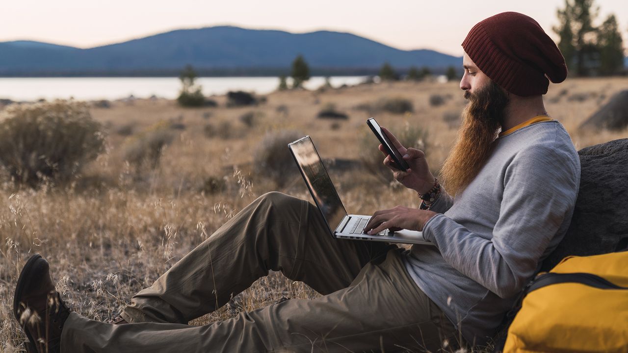 A man using a phone and a laptop out in the wilderness