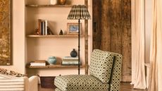 A green patterned cushioned armless chair, in a warm-toned limewashed living room. In front of a green checked lamp, and a rustic wooden door. 