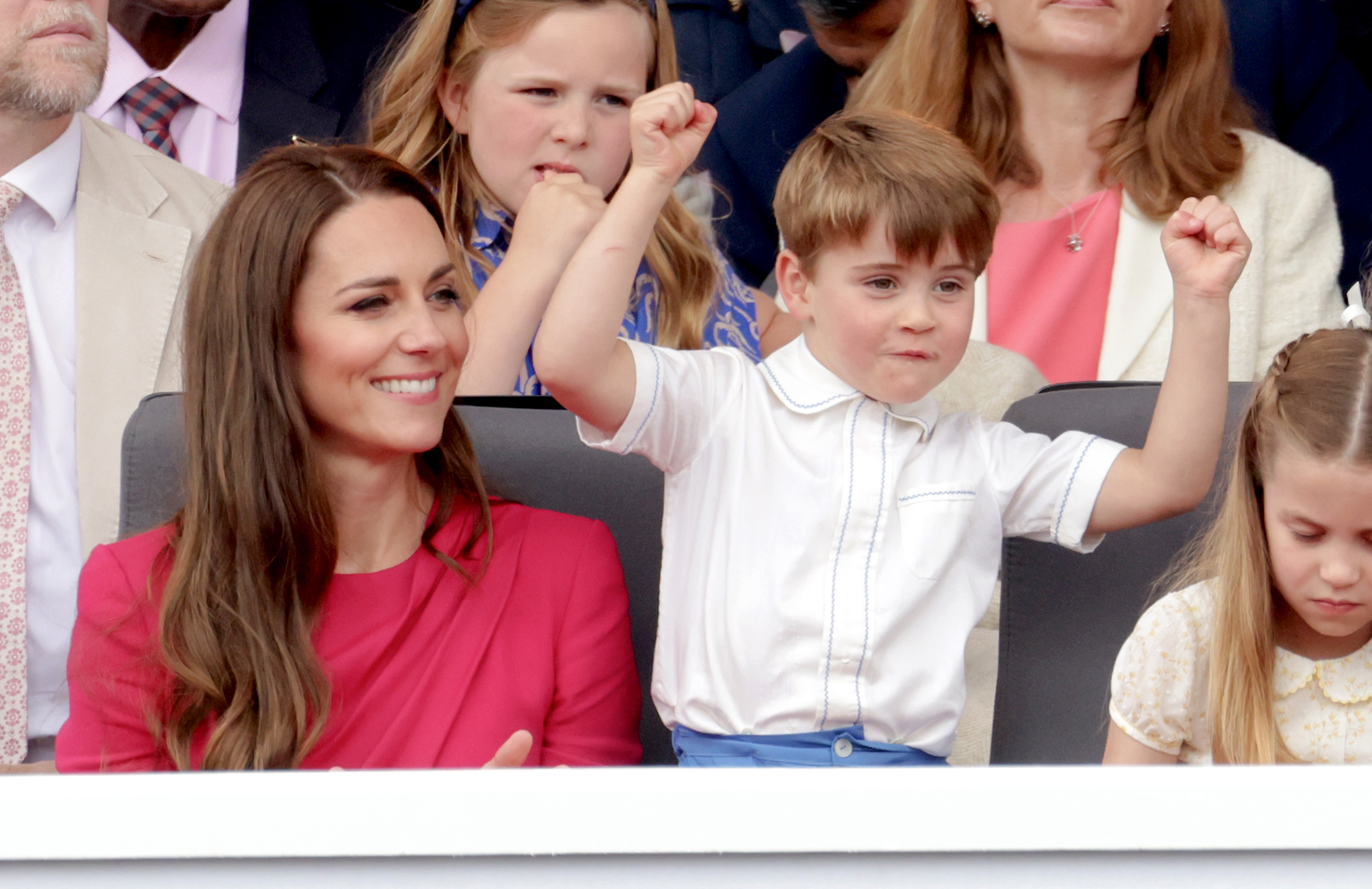 Princess Kate wearing a pink dress sitting next to Prince Louis, who is dancing in his seat