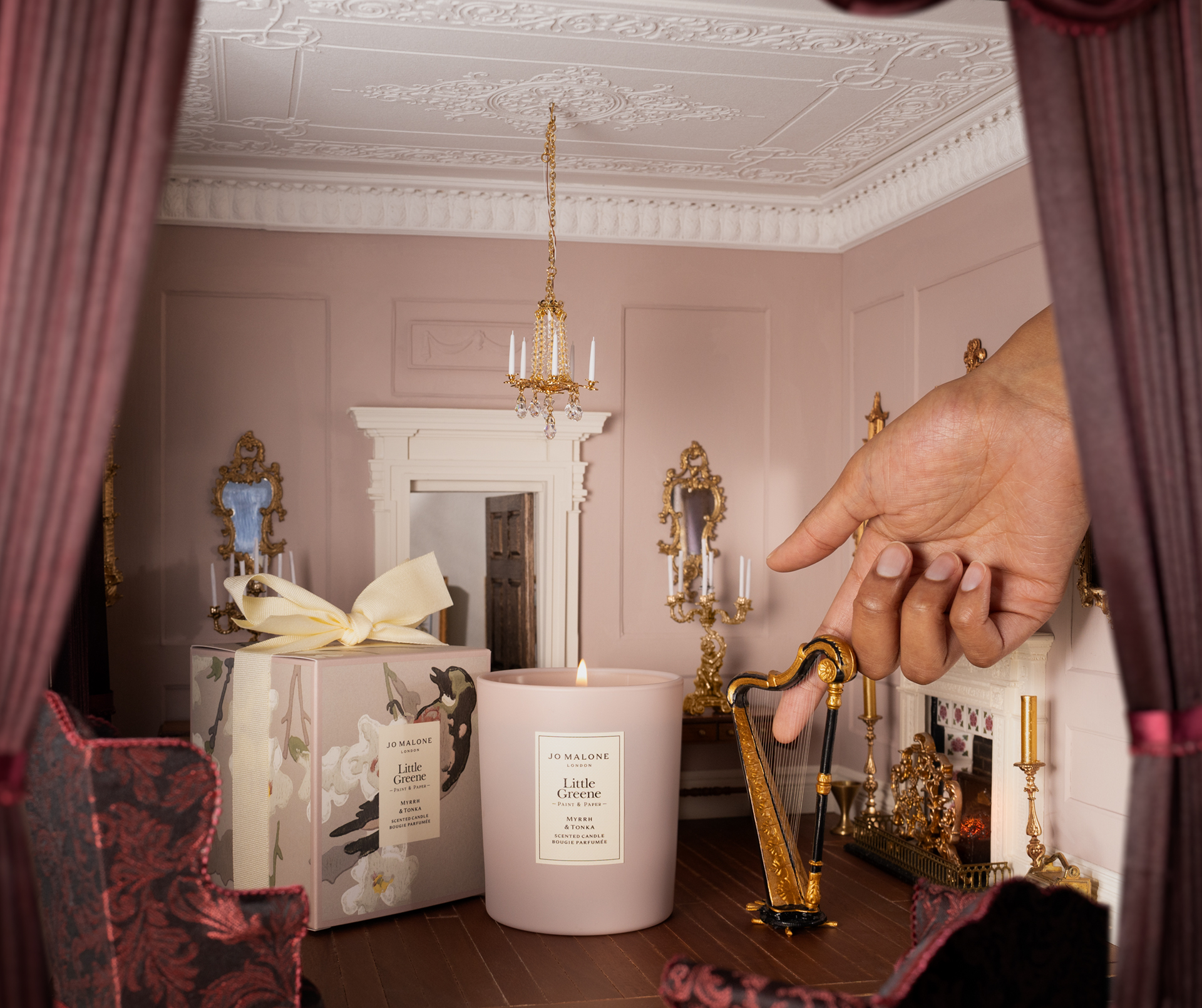 Image of a miniature room that is painted in a blush pink/ purple color with crown molding on the walls and plum curtains in the front. There is a hand reaching in from the side, holding onto a miniature harp. There is a regular-sized candle that's in the middle of the room.