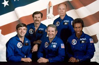 five men in blue flight suits stand in front of an american flag