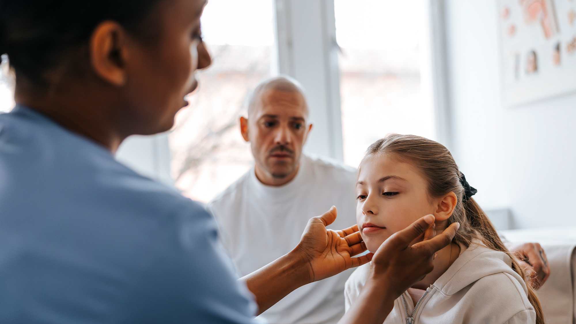 A nurse wearing blue scrubs feels under a small girl's chin while her father watches behind her