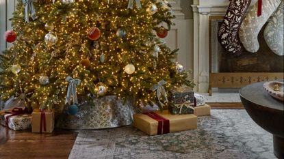 Festive, brightly lit Christmas tree decorated with a brass tree skirt pictured in front of a fireplace located inside of a cozy holiday living room