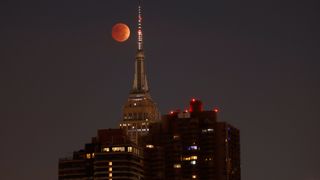 red moon behind tall buildings during total lunar eclipse.