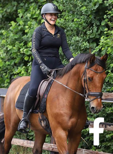 A smiling female horse rider dressed in black trots towards the camera alongside an arena fence with a tall hedge behind on a bay horse.