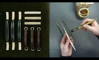 An overview of a crafting table with a mans hands holding a paint brush and a piece of metal on the right and an assortment of brown and black leather bag handles on the left.