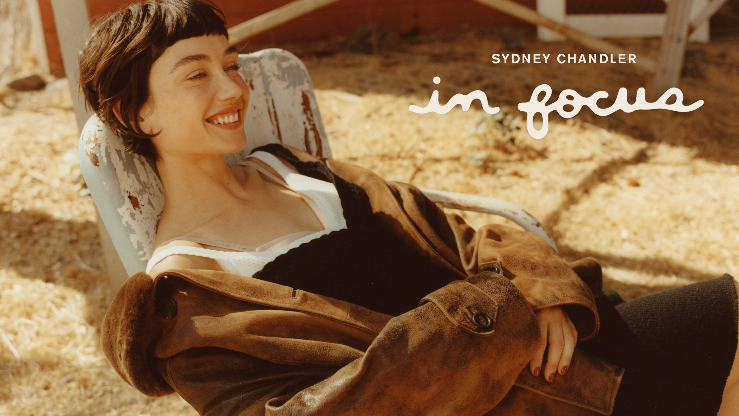 Sydney Chandler for WWW's October In Focus feature. She is sitting in a rusty chair wearing a navy dress and brown leather jacket from Miu Miu. The In Focus logo and her name are written on the image.