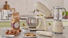 A kitchen countertop with a bag of flour, a crate of eggs, a plate of cookies, a SMEG stand mixer, whisk and blender