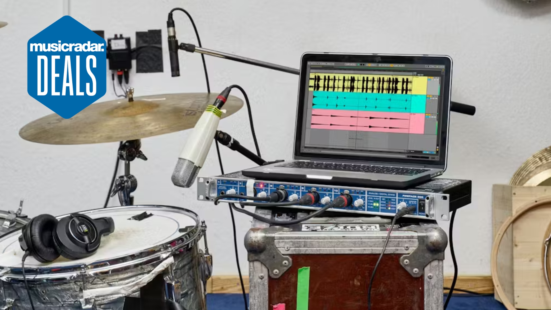 A laptop on top of some music gear with Ableton Live 12 DAW displayed on it. To the left is a drum kit with some headphones and microphones on it.