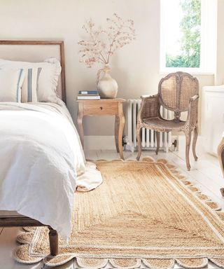 A white bedroom with a scalloped jute rug, a white bed, a chair, and a nightstand