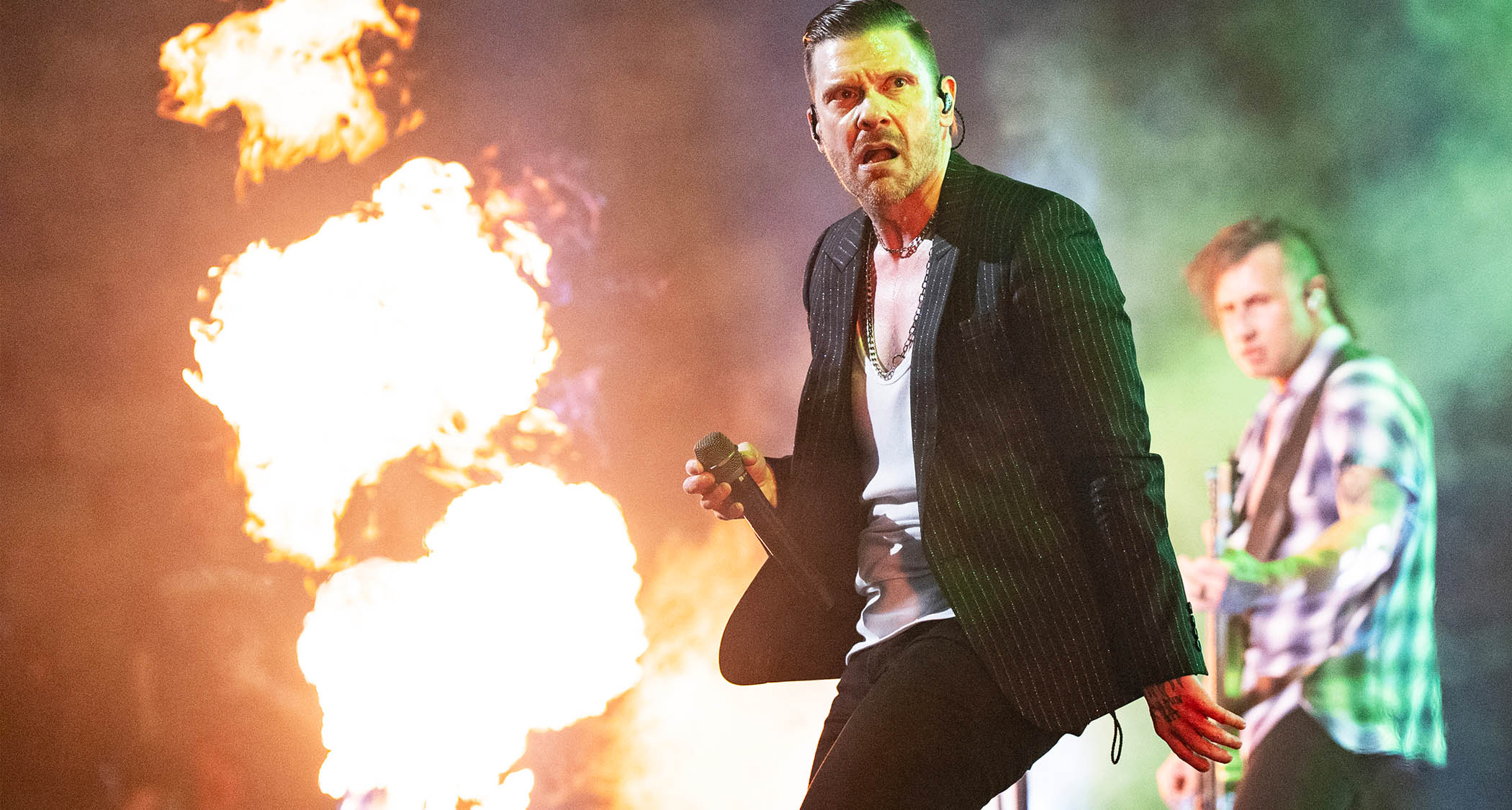 Brent Smith of Shinedown performs on a stage lit with pyro. Bassist Eric Bass is in the background,.