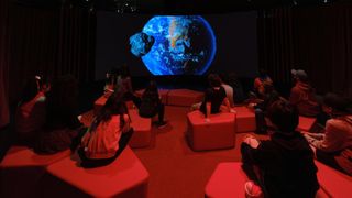 An Earth is projected on a dark screen as children sit on blocks in a dimly red-lit theater