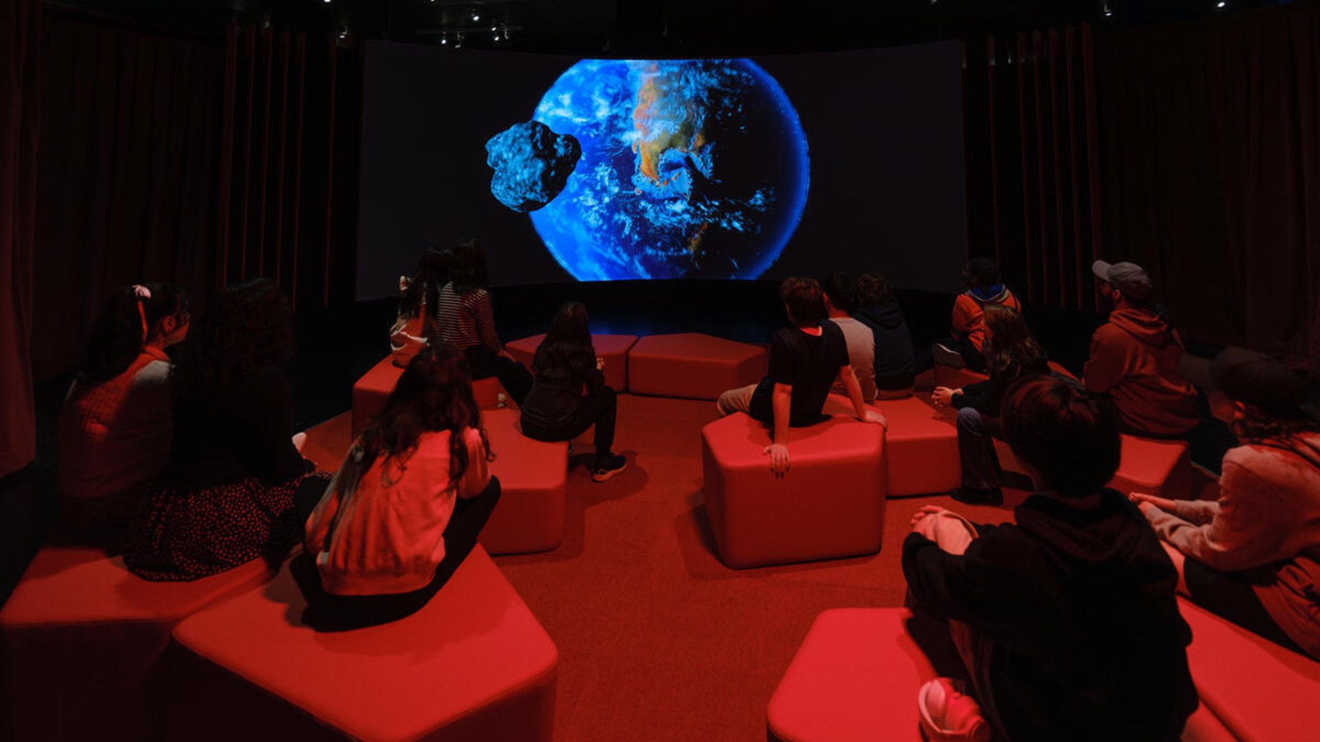 An Earth is projected on a dark screen as children sit on blocks in a dimly red-lit theater