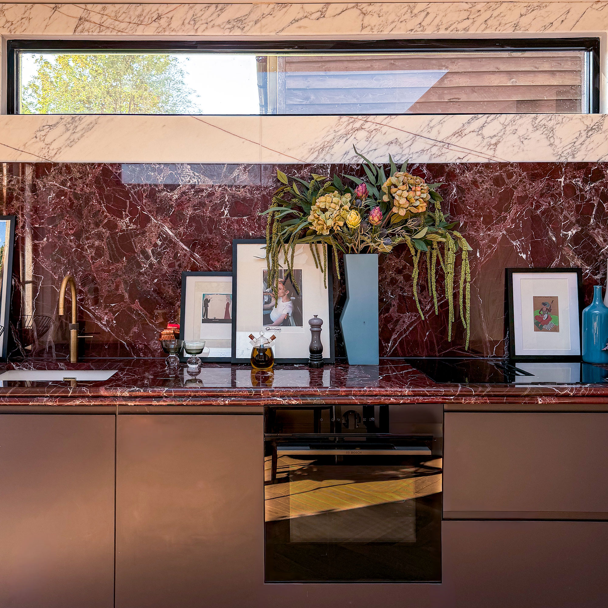 Kitchen with red marble worktops and splashbacks, topped with white marble. Dark red cabinetry. High narrow window. Photographs and vases on countertop