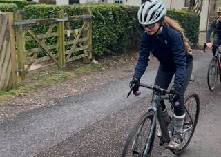 Child in blue with white helmet out the saddle on a gravel bike on the road with gates and hedge in background