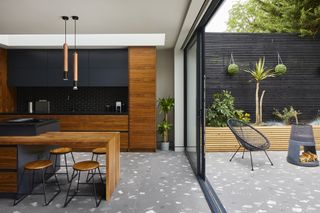 Gray terrazzo kitchen tile ideas flowing out from a modern black and wooden kitchen to a garden with firepit.