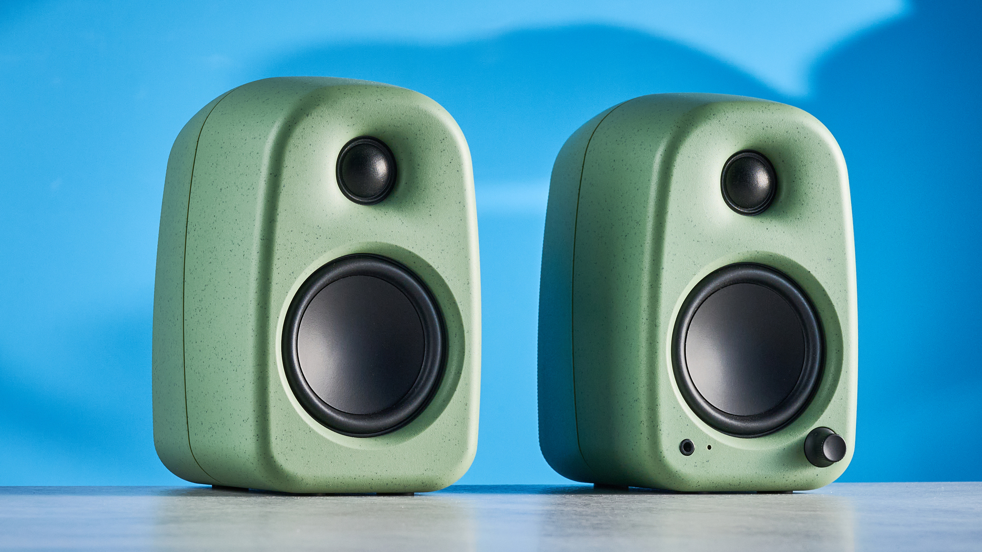 Kanto UKI bookshelf speakers in sage photographed in front of a blue background